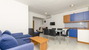 Un salon moderne avec un canapé bleu, une table basse en bois et un coin repas comprenant une table et des chaises. On aperçoit une cuisine avec des meubles bleus et un téléviseur à l’arrière-plan.