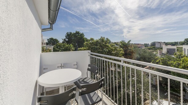 Un balcon avec une table et des chaises blanches, une rambarde en métal et une vue sur les bâtiments et la végétation environnants.