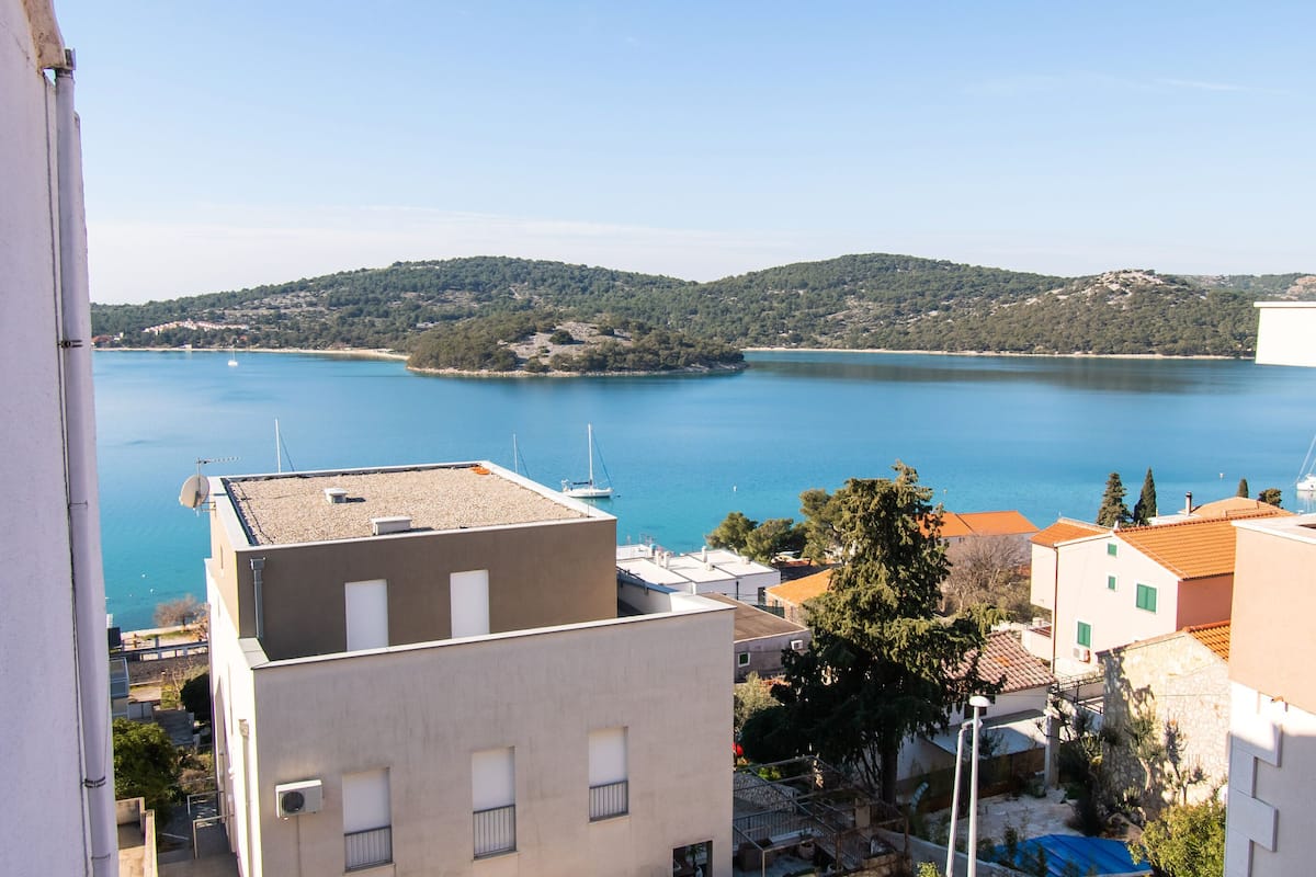 A coastal town with buildings overlooking a calm sea, a small island, and hills in the distance.