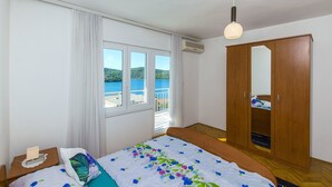 Een slaapkamer met een bed, nachtkastje, kast, raam met gordijnen en uitzicht op zee.
