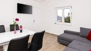 Een moderne woonkamer met een grijze bank, een witte eettafel met stoelen, een aan de muur bevestigde televisie en een raam met uitzicht op heuvels.