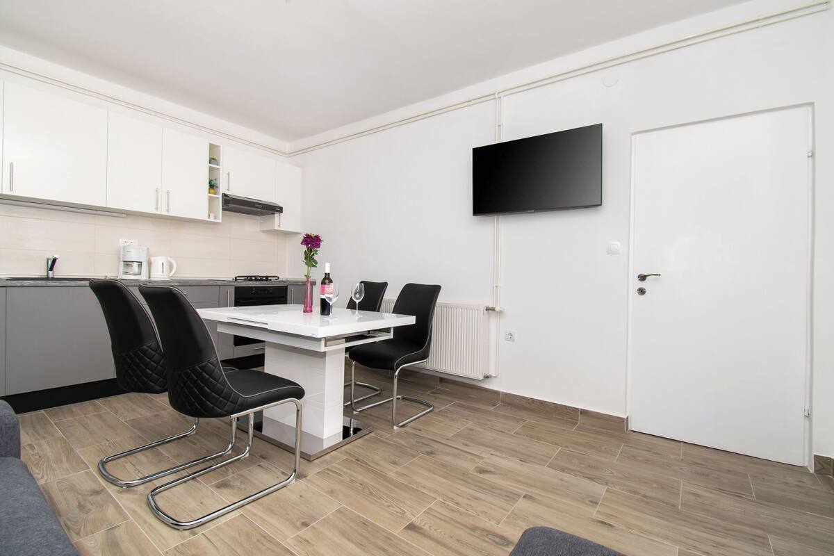 A modern kitchen with white cabinets, a white dining table with black chairs, a coffee maker, and a flower vase.