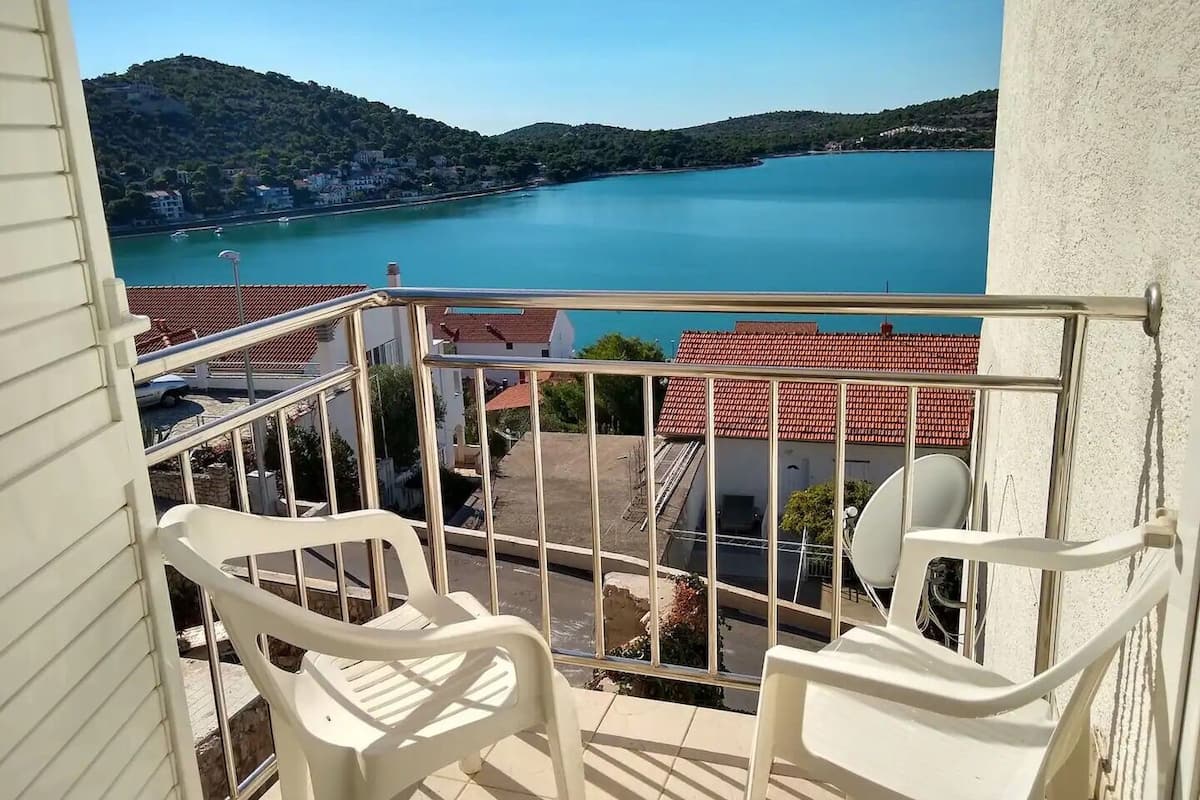 A balcony with white plastic chairs, a metal railing, and a view of a turquoise lake surrounded by hills.