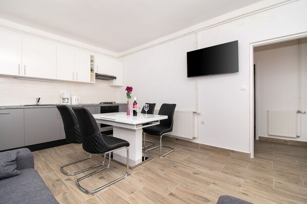 Una cocina moderna con armarios blancos, una mesa de comedor con sillas, un televisor de pantalla plana montado en la pared y un sofá gris.