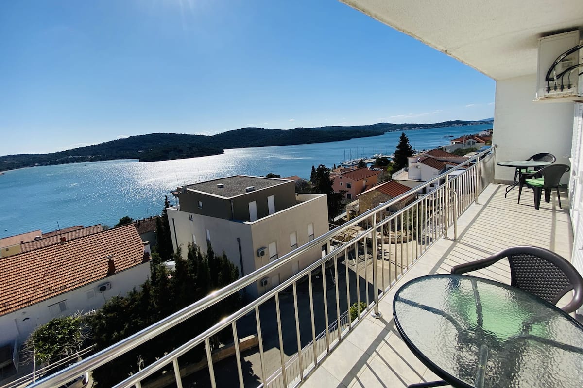 A balcony with a glass table and chairs overlooking a lake with boats, surrounded by buildings with red-tiled roofs.
