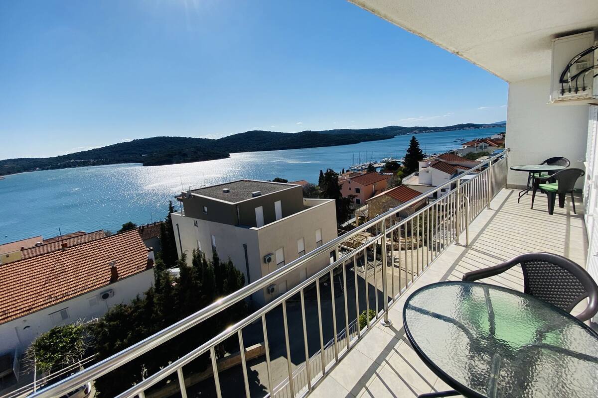 A balcony with a glass table and chairs overlooking a lake with boats, surrounded by buildings with red-tiled roofs.