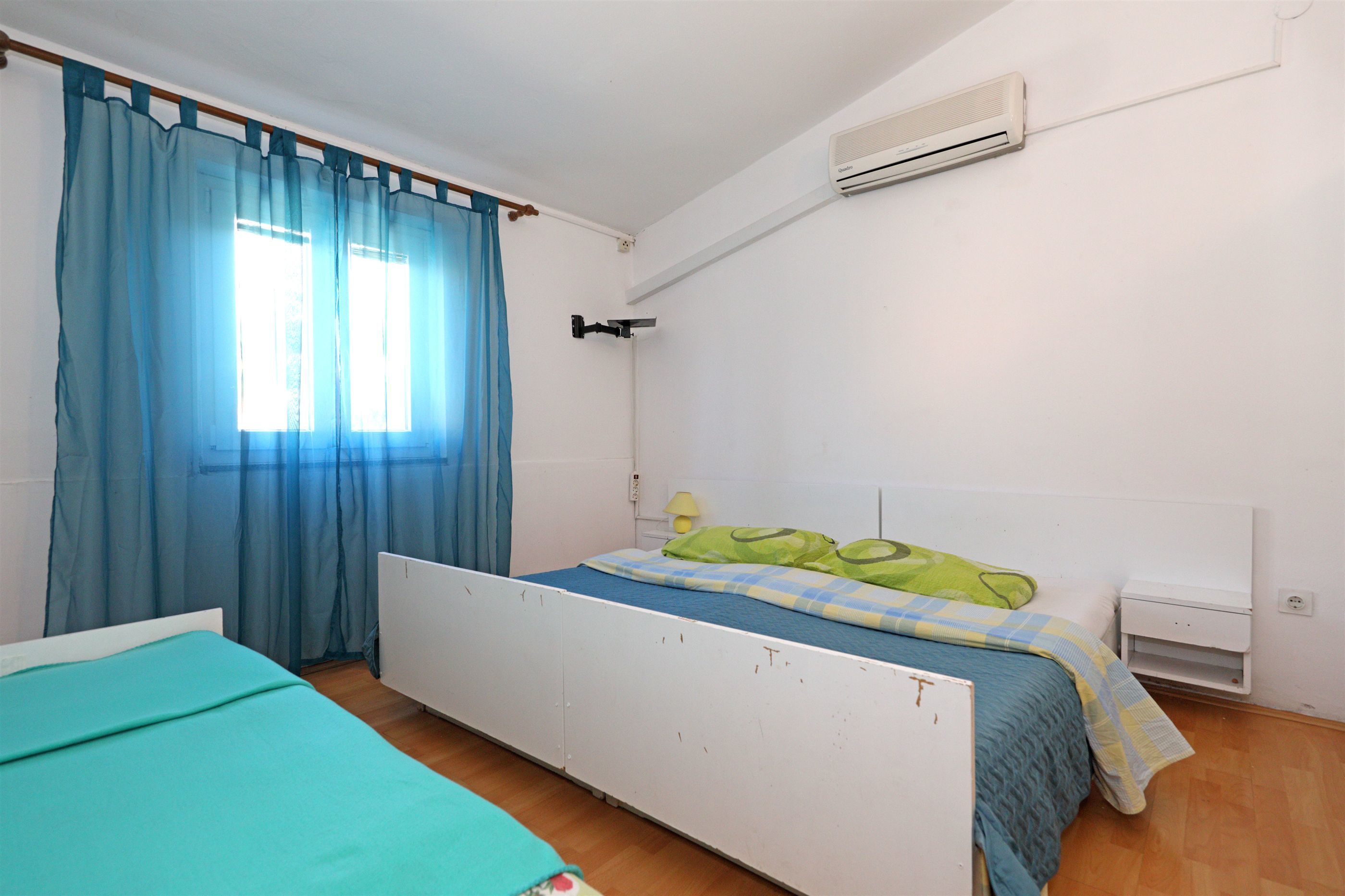 Une chambre avec un lit simple, une table de chevet, un climatiseur fixé au mur et une fenêtre avec des rideaux bleus.