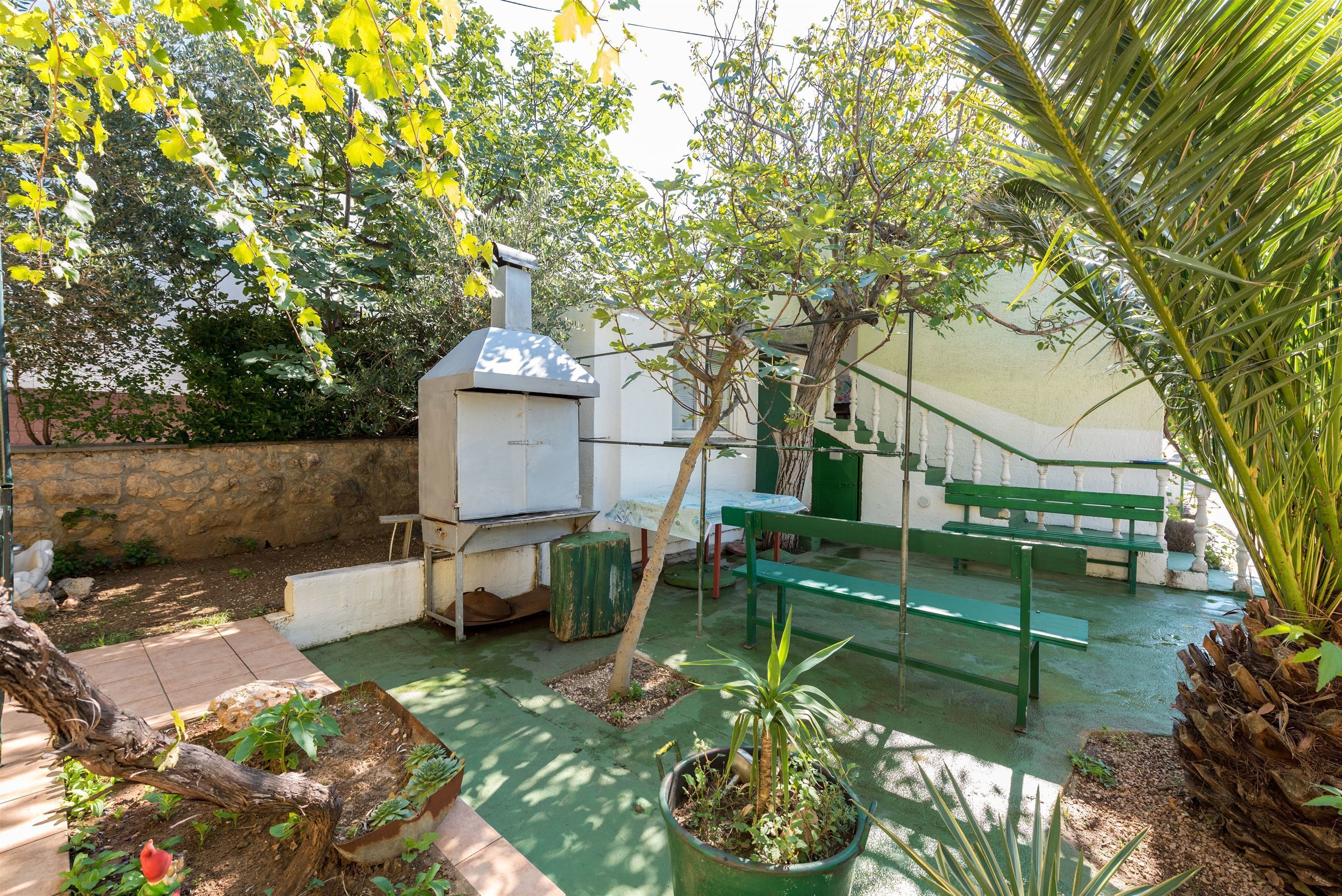 A patio area with a barbecue grill, green benches, potted plants, and a stone wall.