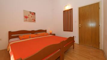 A bedroom with a wooden bed, an orange bedspread, a nightstand with a lamp, a wooden wardrobe, and a wall-mounted light fixture.