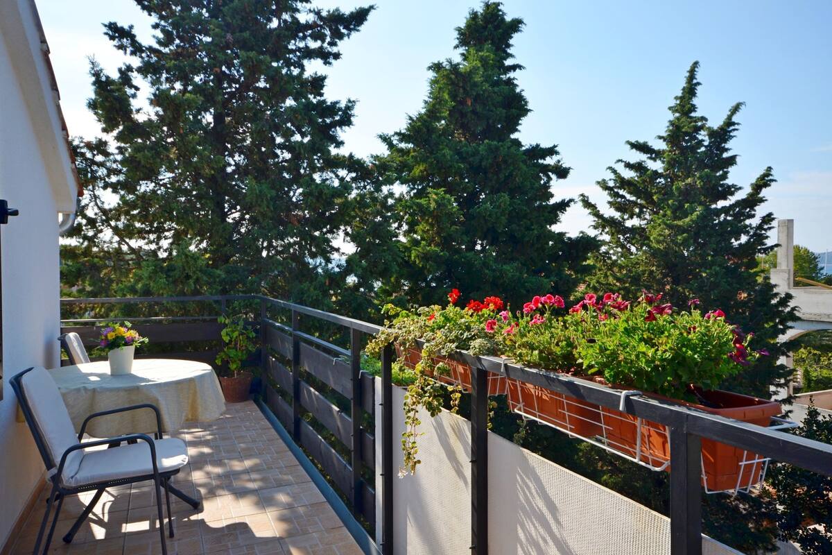 Ein Balkon mit Tisch und Stühlen, Blumentöpfen und Blick auf hohe Bäume.
