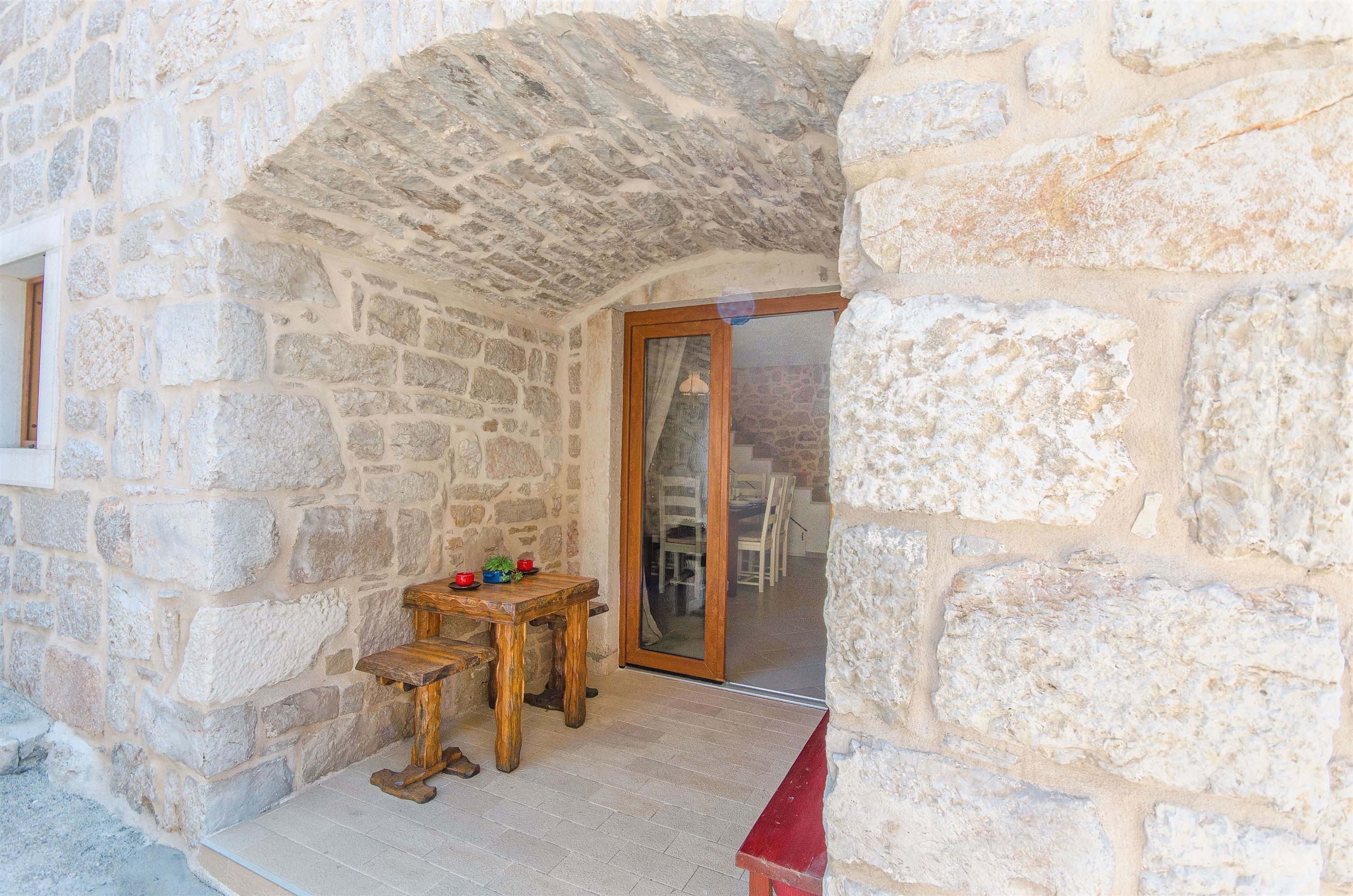 A stone-walled room with a wooden table and chairs, a wooden bench, and a red bench.