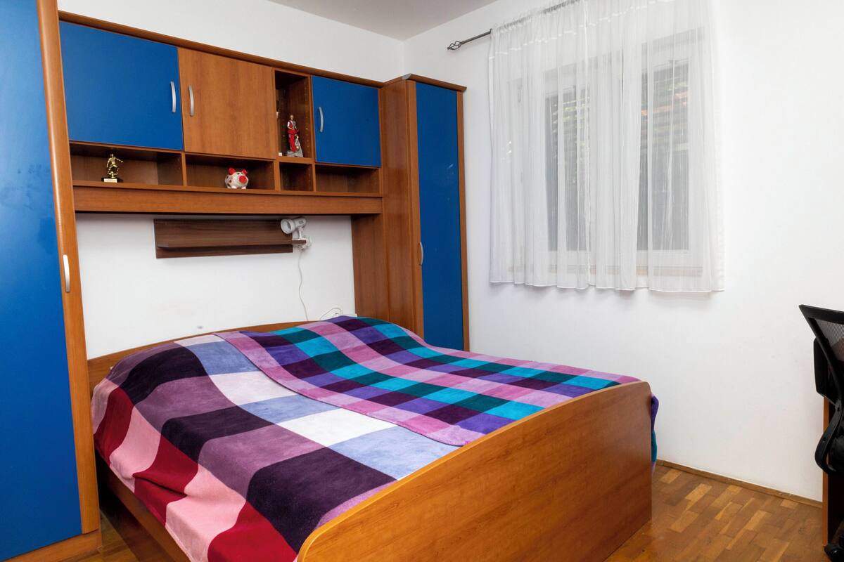 Une chambre avec un lit en bois, une couverture à carreaux aux couleurs vives, une armoire bleue et en bois, et une fenêtre ornée de rideaux légers.