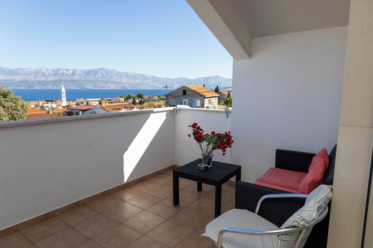 Un balcon avec vue sur la mer et les montagnes, une chaise en osier, une petite table avec un vase de fleurs et un coussin rouge.