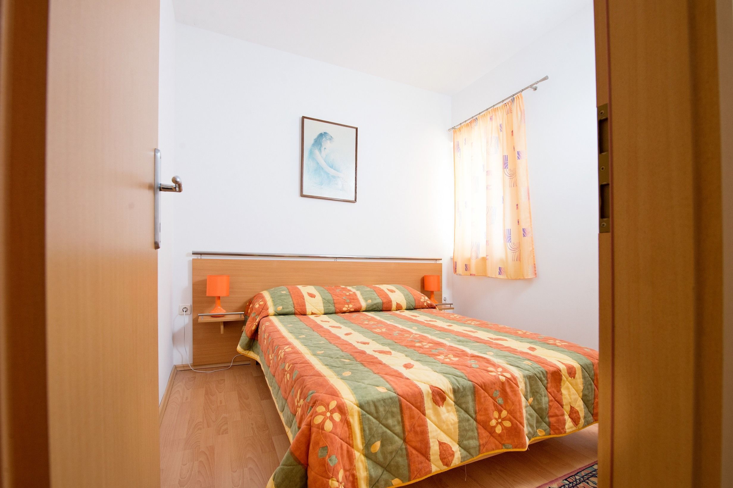 A bedroom with a wooden bed, a nightstand, a lamp, a framed picture on the wall, and a window with curtains.