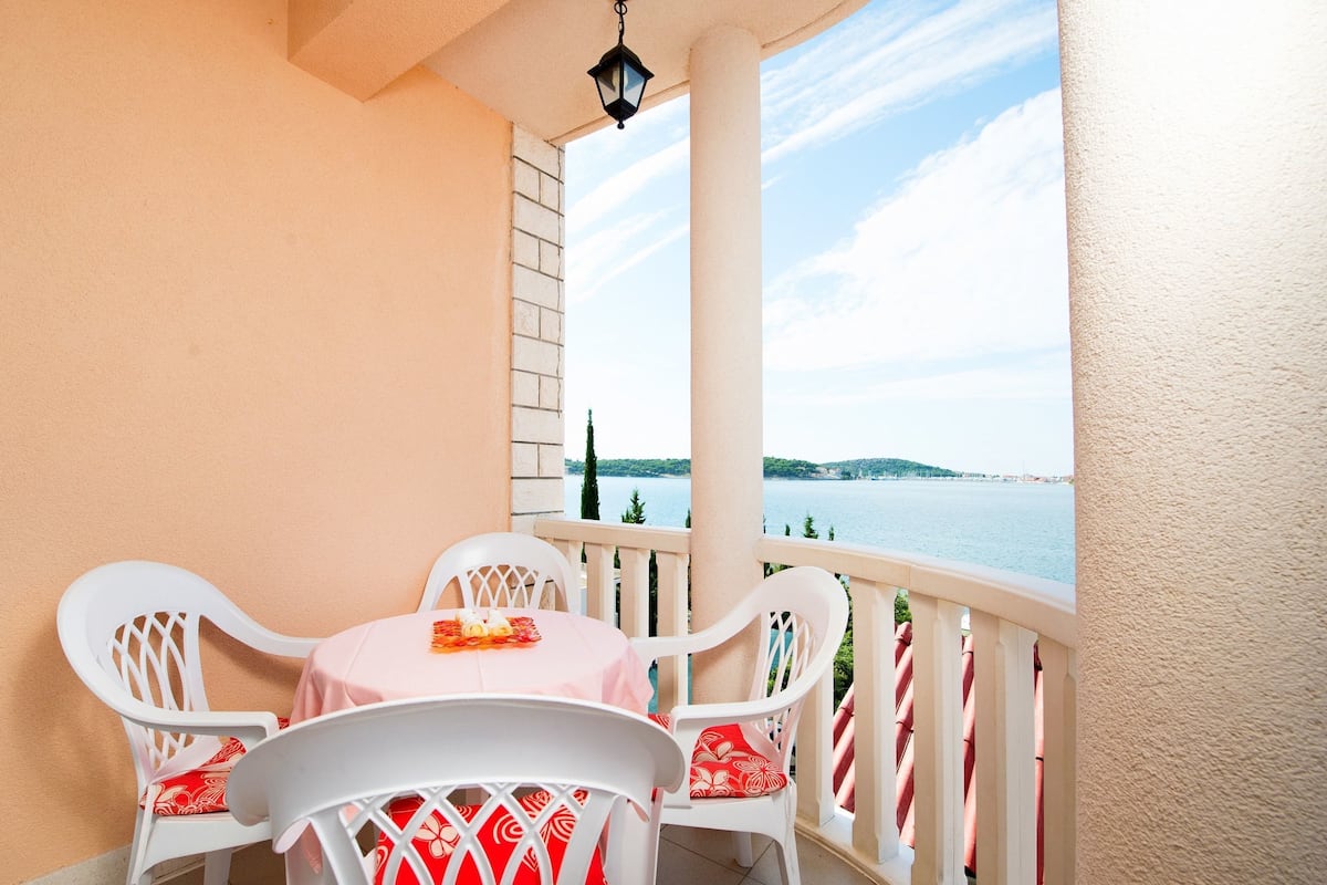 Ein Balkon mit Tisch und Stühlen, mit Blick auf ein Gewässer und eine entfernte Küstenlinie.