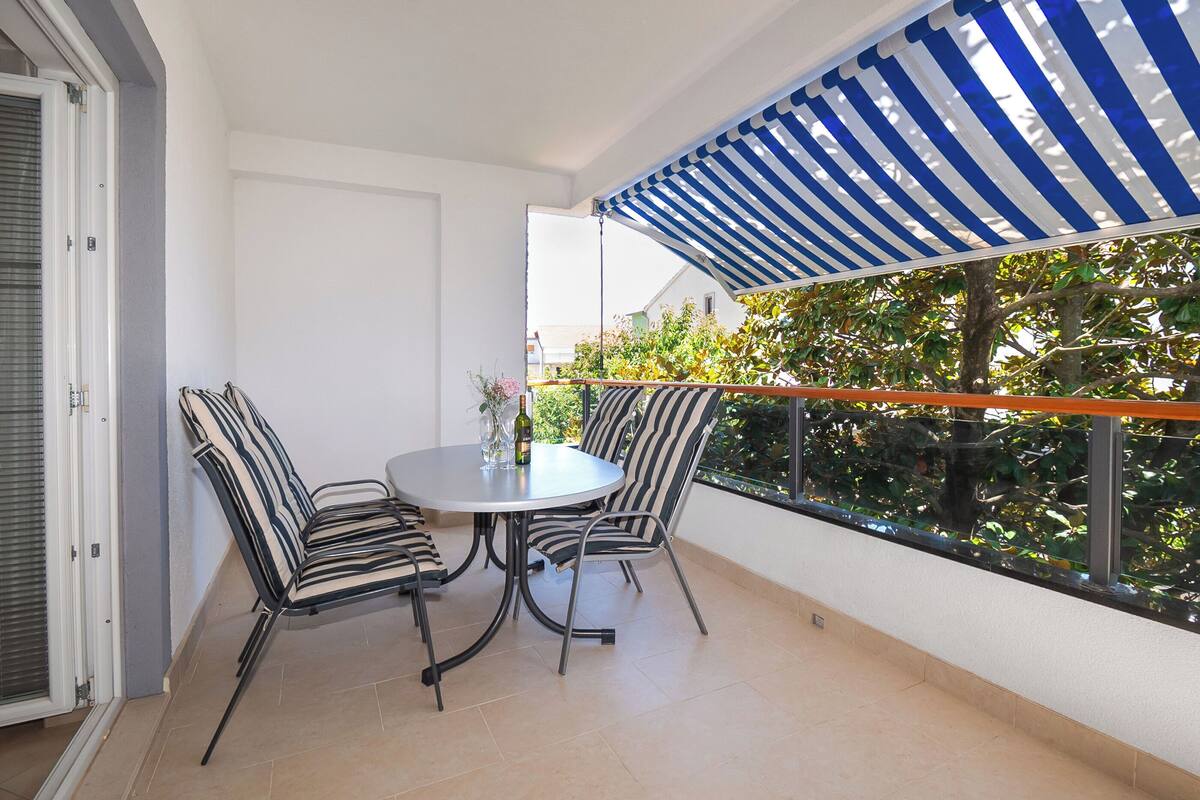 A balcony with a round table and striped chairs, a blue and white striped awning, and a view of greenery.