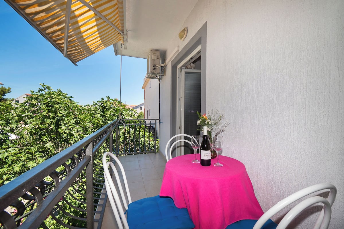 A balcony with a table set for two, featuring a bottle of wine and glasses, and a chair with a blue cushion.