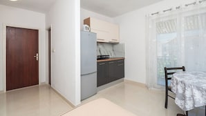 Cocina compacta con refrigerador, estufa y alacenas, una mesa de comedor con una silla, y un ventanal con persianas.