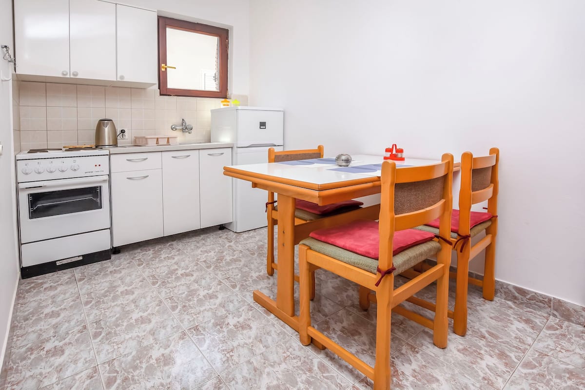 A kitchen with white cabinets, a stove, and a refrigerator. There is a dining table with chairs and a window above the sink.