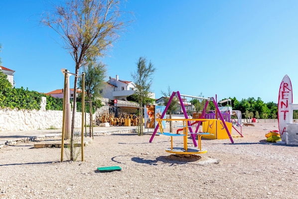 A playground with swings and a slide, surrounded by a gravel area and a white wall.