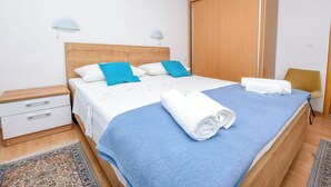 En pænt redt seng med et blåt tæppe og hvide puder, et trænatbord med en skuffe, en garderobe og et lille bord med en stol.