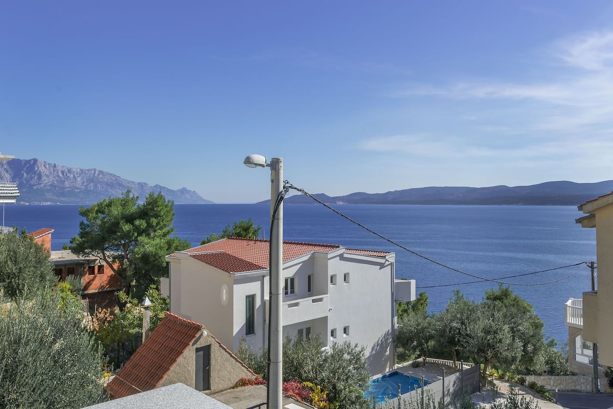 A coastal residential area with white buildings, a swimming pool, and a view of the sea and mountains.