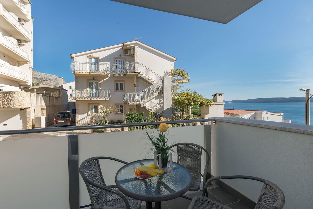 A balcony with wicker chairs and a table, overlooking a coastal town with buildings and a clear sky.