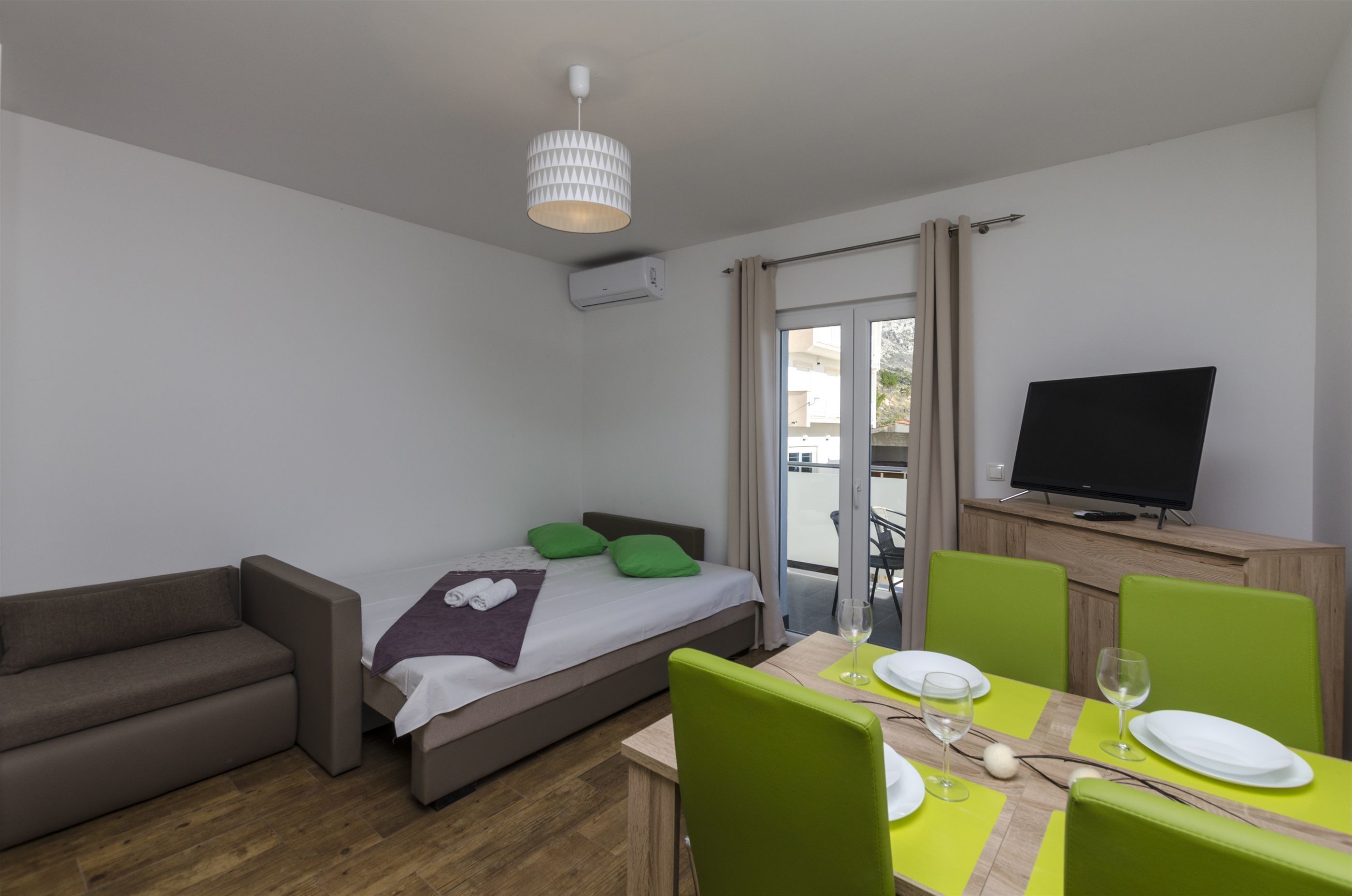 A modern hotel room with a bed, a sofa, a dining table with chairs, a television, and a balcony with curtains.