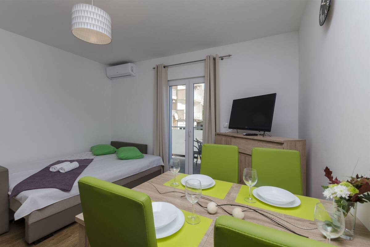 A modern hotel room with a bed, a dining area with green chairs and a table set for two, a flat-screen TV, and a balcony with a view.