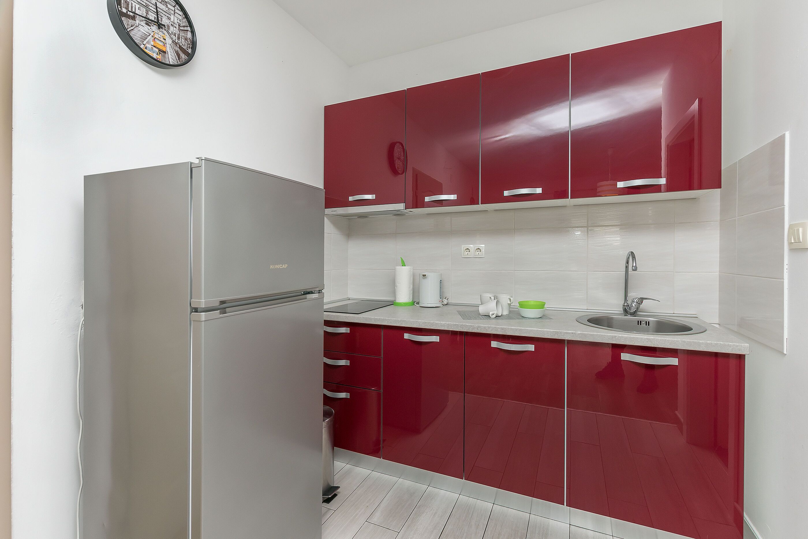 A kitchen with a refrigerator, red cabinets, a sink, and a clock on the wall.