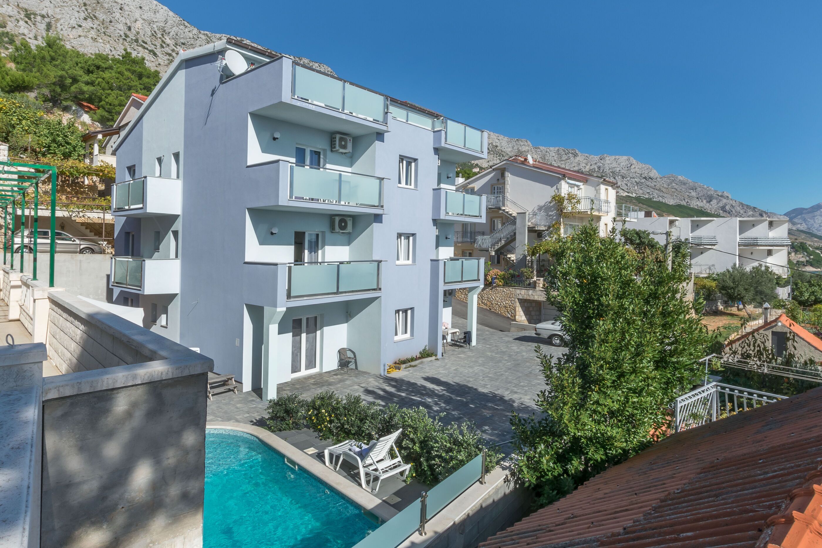 A modern multi-story building with balconies, a swimming pool, and a mountainous backdrop.