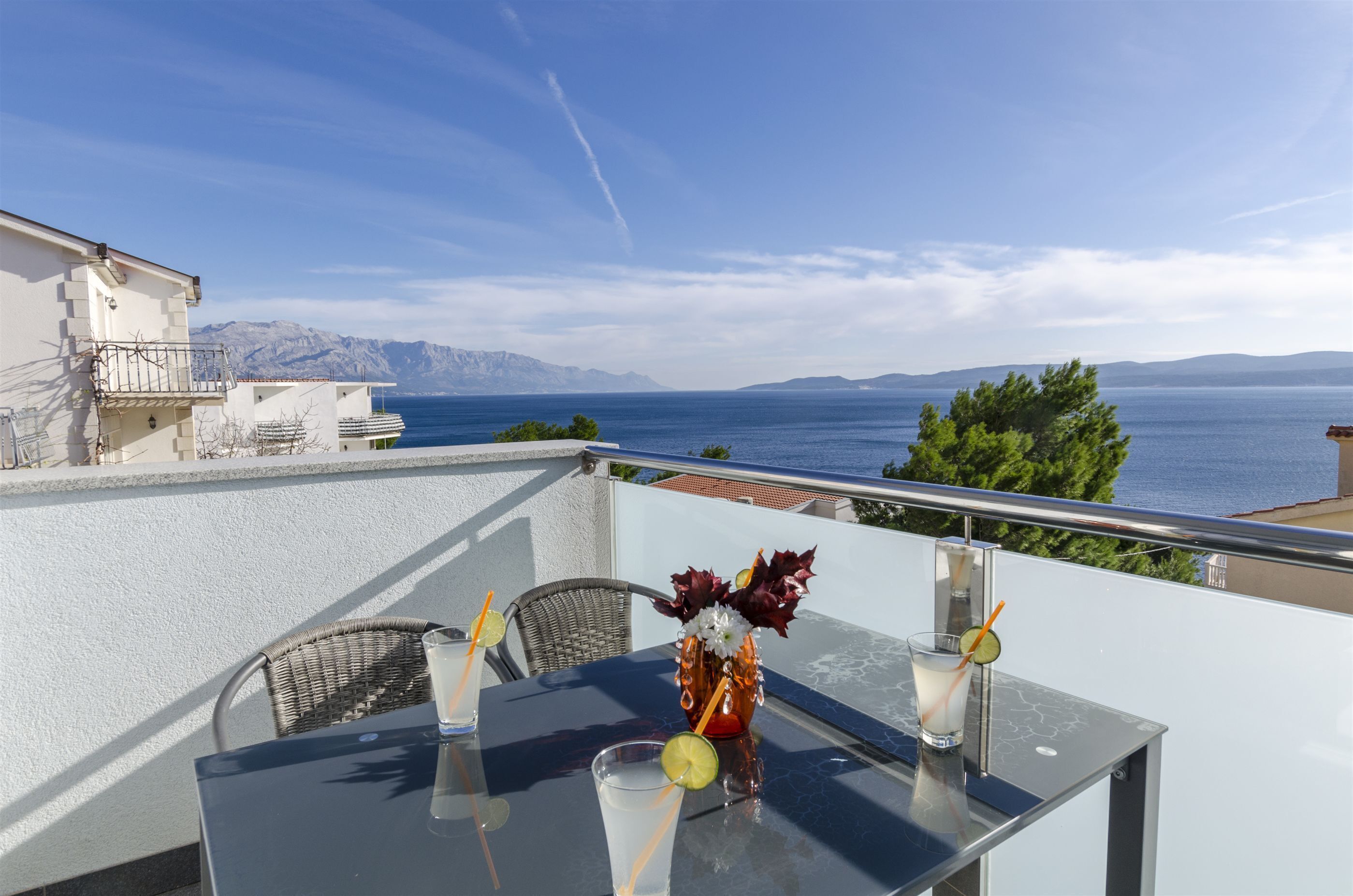 A balcony with a table set for two, a vase with flowers, and a clear view of the sea and mountains.