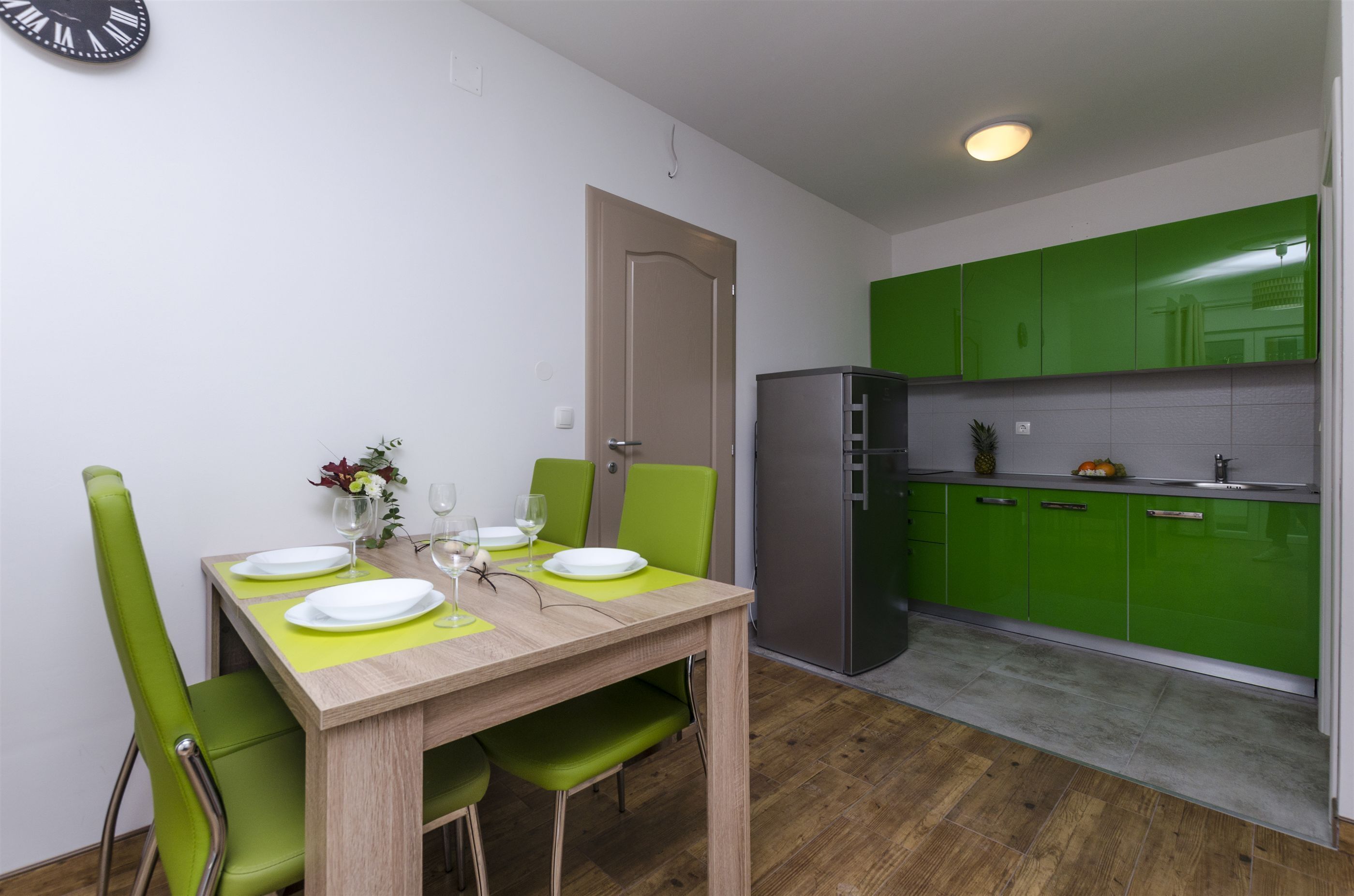 Een compacte keuken met groene kastjes, een houten eettafel gedekt met borden en glazen, en een klok aan de muur.