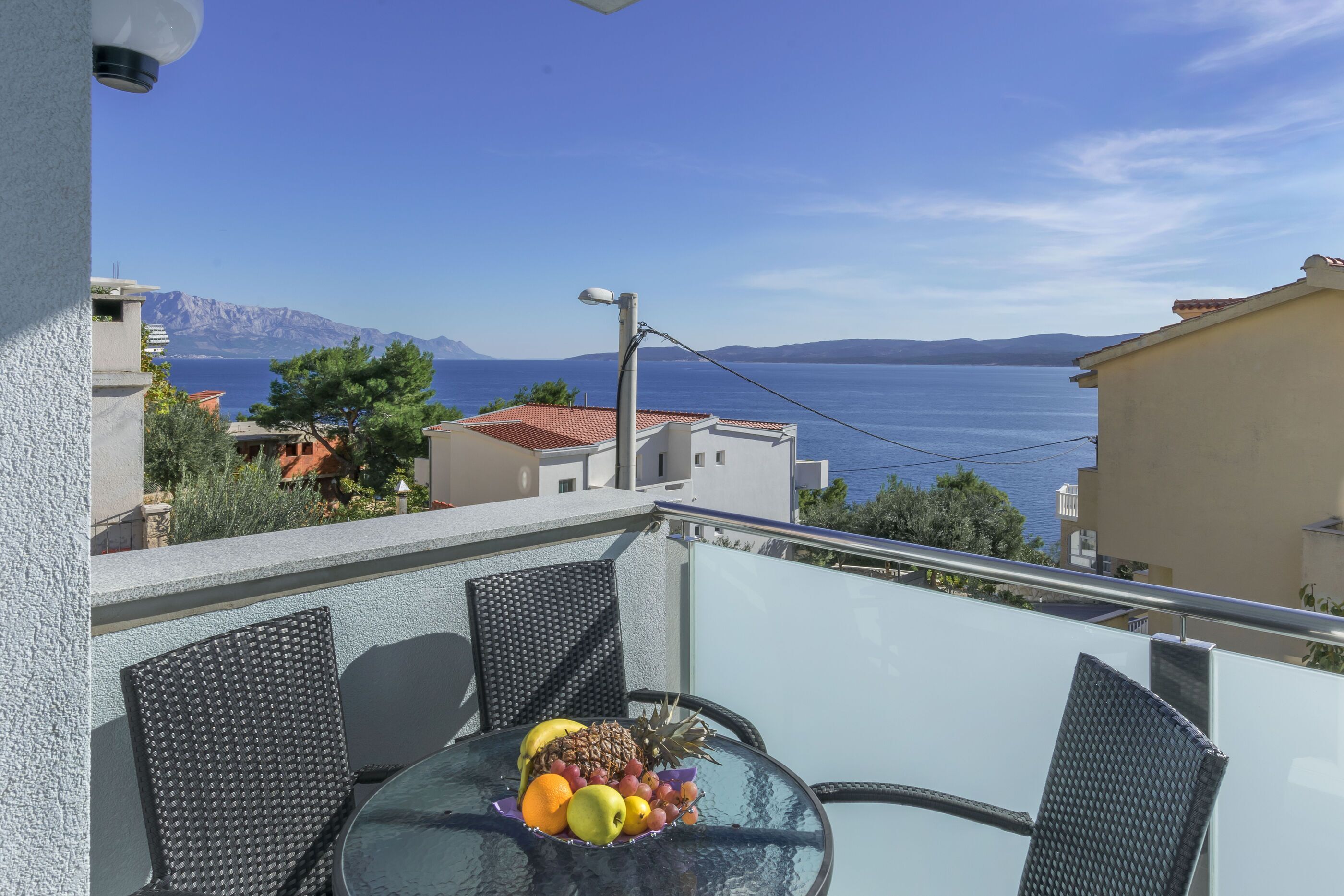 A balcony with a glass table, wicker chairs, and a fruit bowl. The view includes a body of water and distant mountains.
