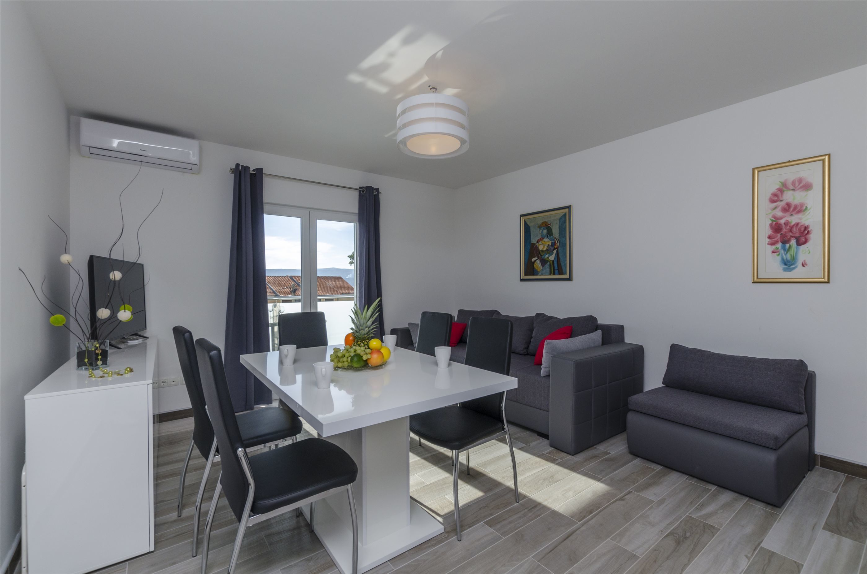 Uma sala de estar moderna com área de jantar, sofás cinza, uma mesa branca e uma luminária no teto.