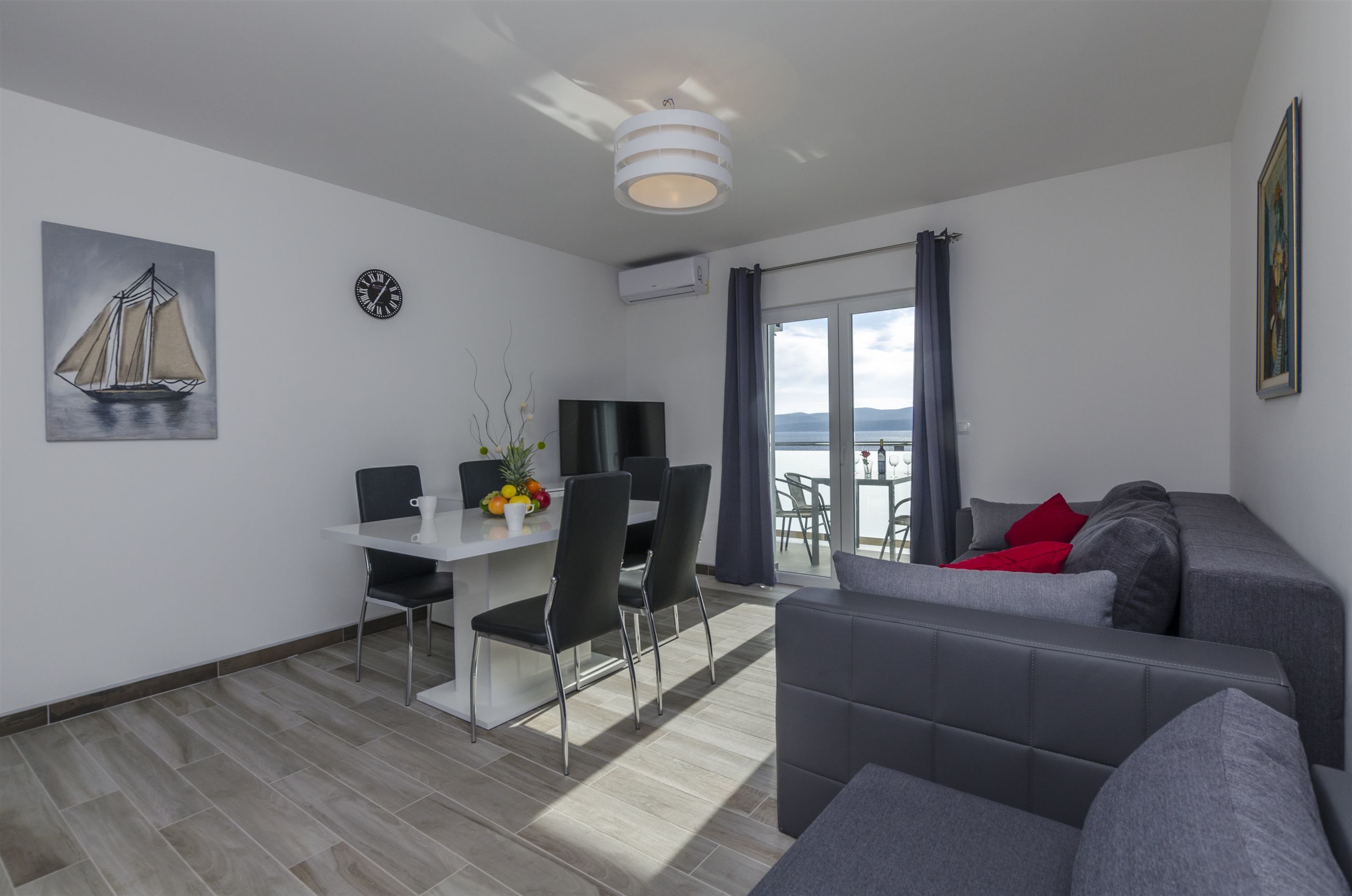Una sala moderna con un sofá gris, una mesa de comedor con sillas, un televisor de pantalla plana y un cuadro de un velero.