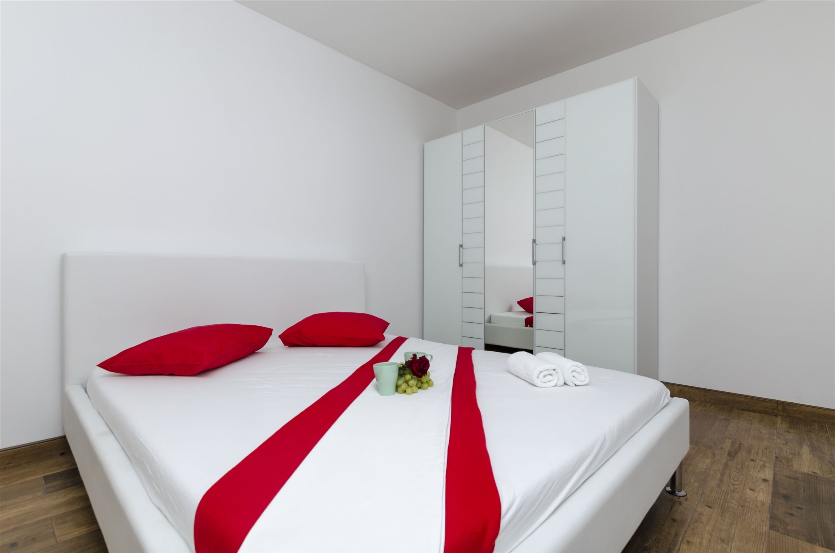 A hotel room with a bed, red pillows, a white bedspread, a red runner, a small table with a vase of flowers, and a white wardrobe with a mirror.