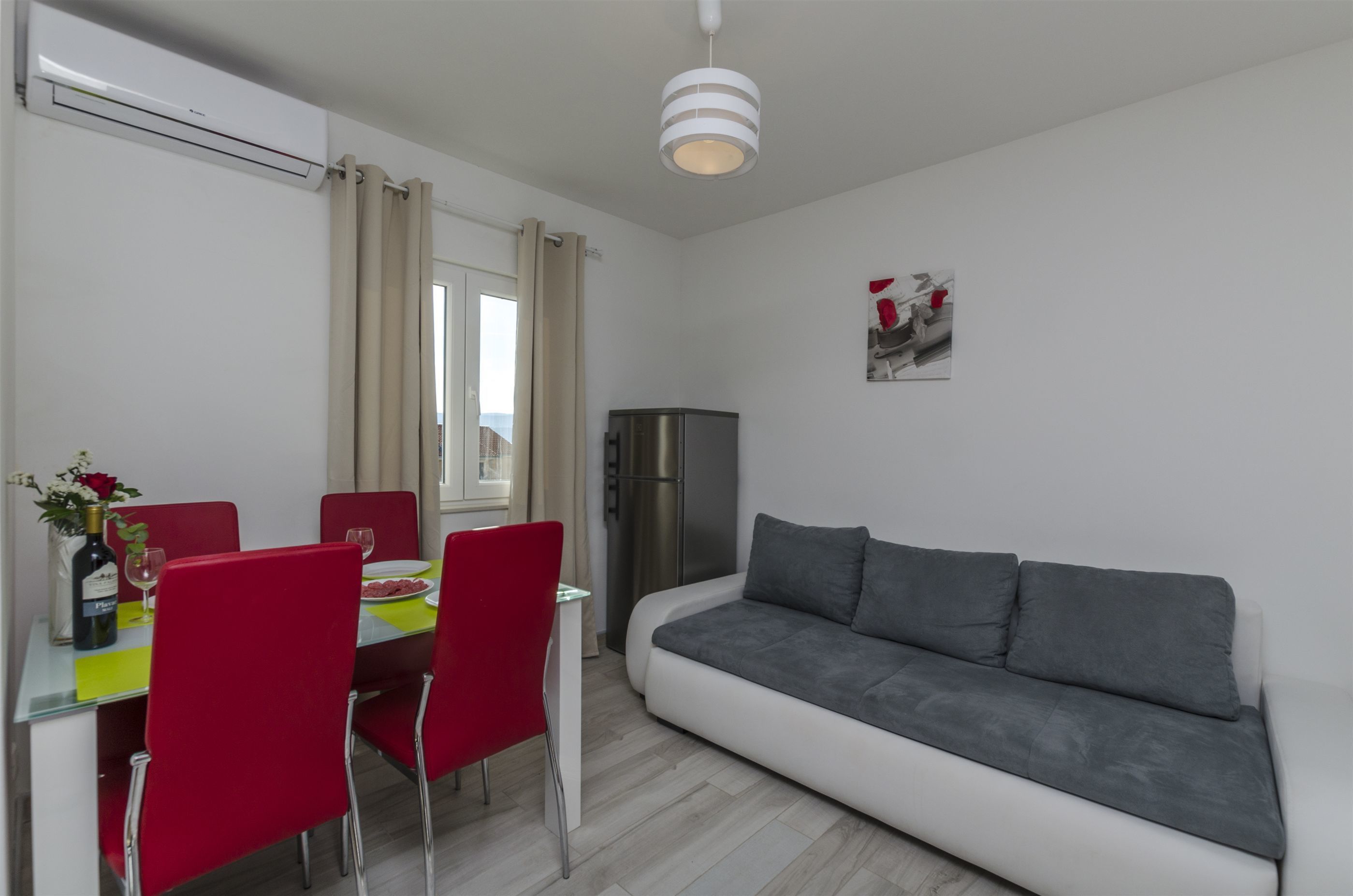 Een moderne woonkamer met een grijze bank, een eettafel met rode stoelen en een roestvrijstalen koelkast.