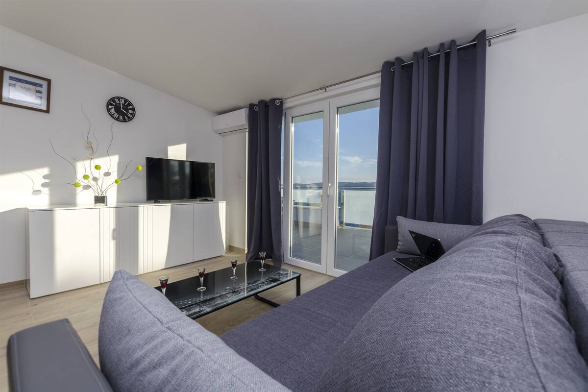 A modern living room with a grey sofa, a flat-screen TV, a dining area with a white cabinet, and a balcony with curtains.