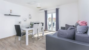 A modern living room with a grey sofa, a dining table with chairs, and a large window with curtains. - Apartments Lorenzo (Lokva Rogoznica)