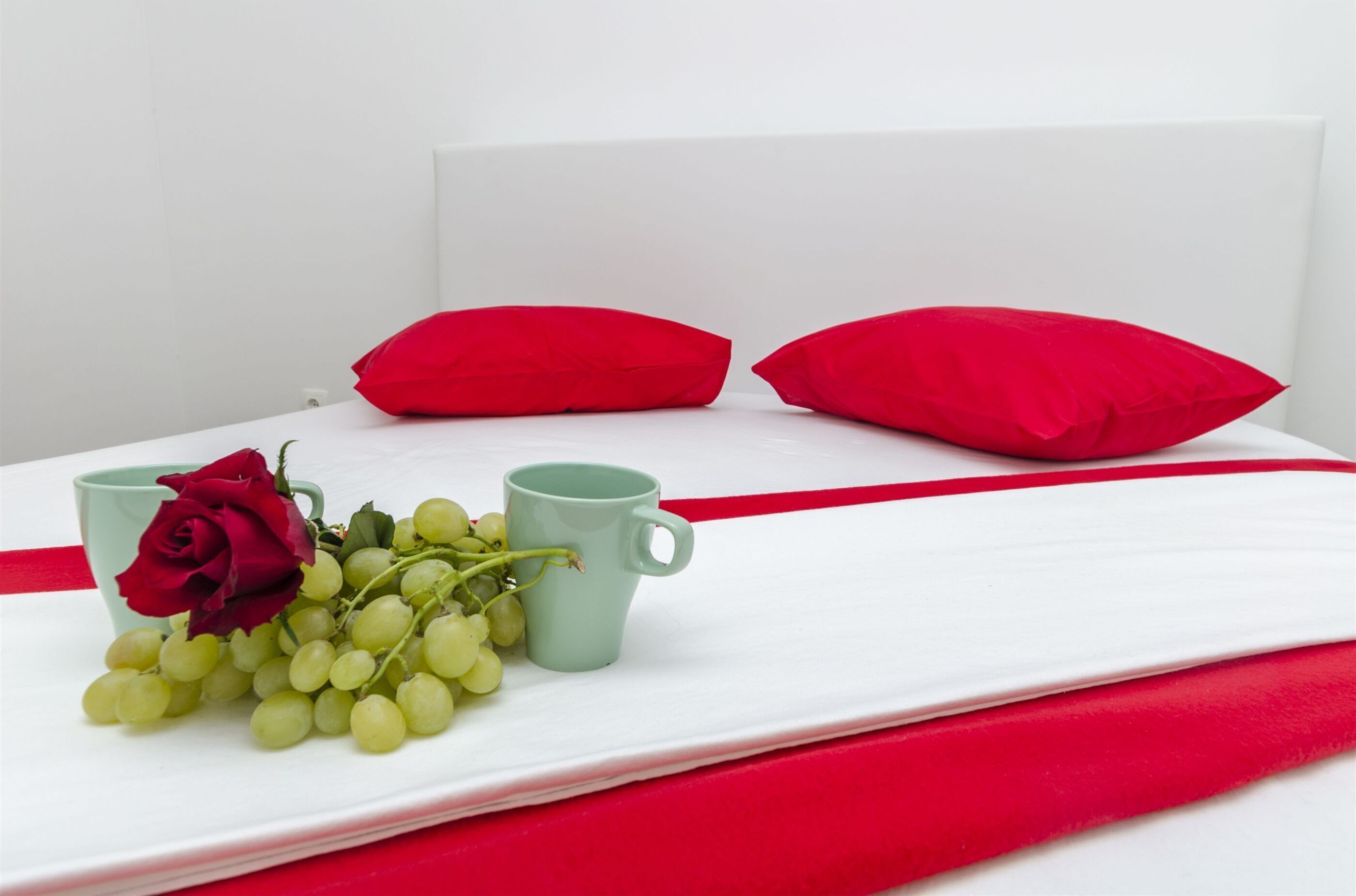 Cama bem arrumada com lençol branco, almofadas vermelhas, uma caneca verde e um buquê de uvas verdes e uma rosa vermelha.