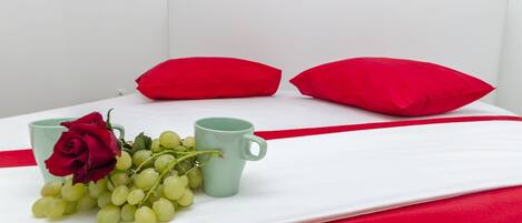A neatly made bed with a white sheet, red pillows, a green mug, and a bouquet of green grapes and a red rose.
