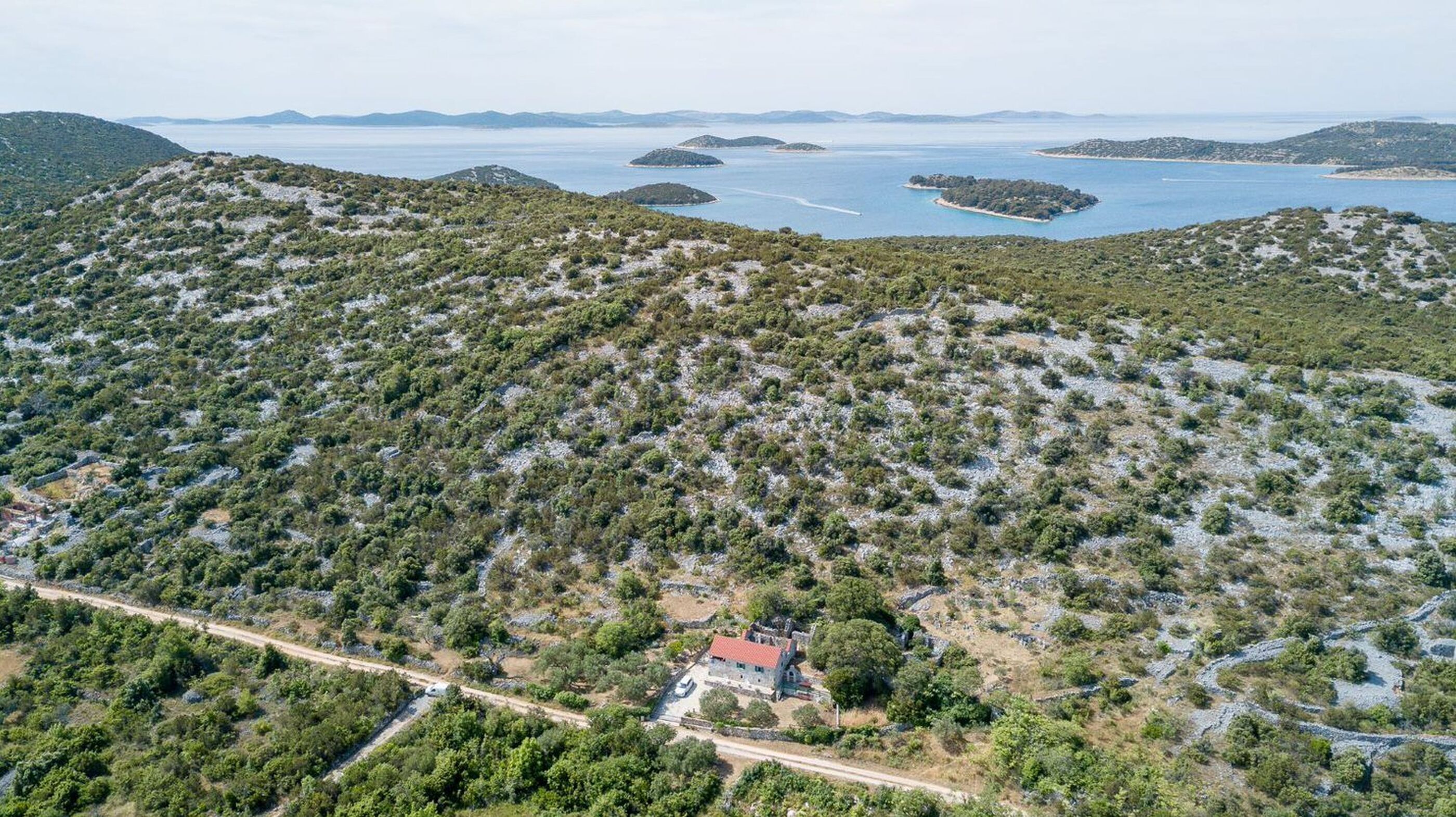 An aerial view of a mountainous area with scattered vegetation and a small building with a red roof near a road.