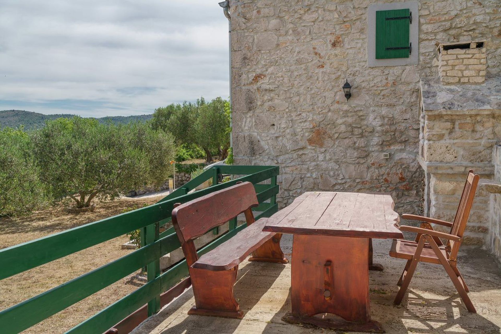 An outdoor seating area with a wooden table and benches, a stone wall, and green shutters.