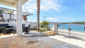 A patio with stone flooring, outdoor furniture, a palm tree, and a view of the sea through a metal fence. - Apartment Iryna (Rogoznica)