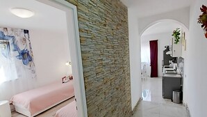 A modern interior with a stone accent wall, a bed with a pink blanket, a kitchenette, and a bathroom visible through an archway. - Apartment Iryna (Rogoznica)