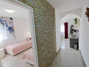 Un intérieur moderne avec un mur d’accent en pierre, un lit recouvert d’une couverture rose, une kitchenette et une salle de bain visible à travers une arche.