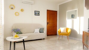A living room with a sofa, a small table with a bowl of fruit and flowers, a yellow armchair, and a wooden door.