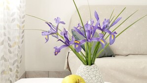A white table with a bowl of green apples and a vase of purple flowers.