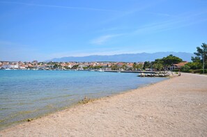 Uma cidade costeira com praia de areia, águas calmas e montanhas ao fundo.